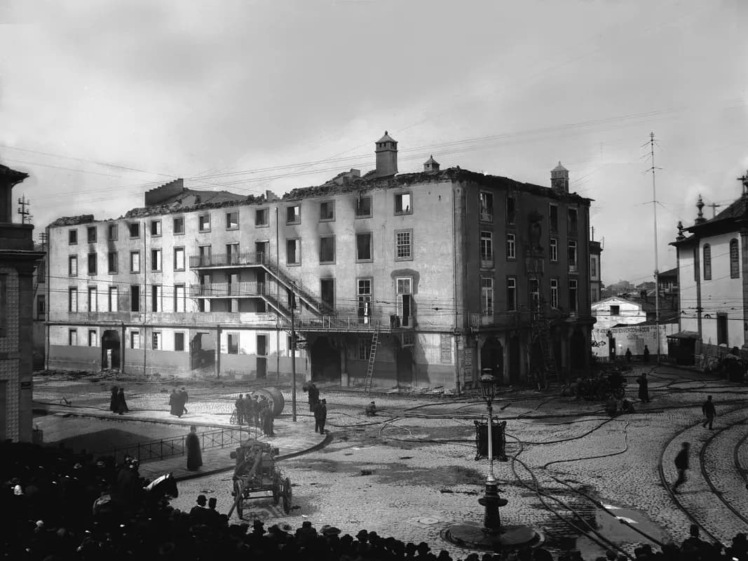 Black-and-white photograph of a long, four-storey building with no roof and blackened, empty windows. Fire hoses snake across the cobbled square in front, a few men stand by a hose reel, and the heads of a crowd fill the bottom of the picture.