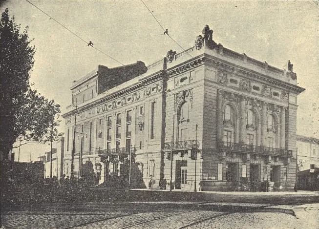 Black-and-white photograph of a large theatre on a street corner, with an ornate upper storey, arched windows and sculpture on the roofline. Tram tracks cross the cobbles in the foreground and a tree stands on the left.