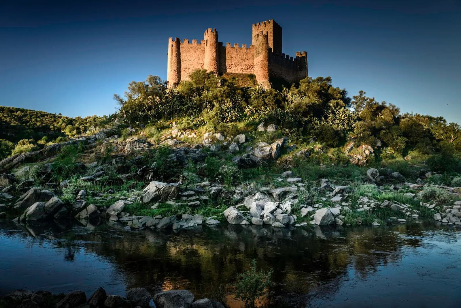 A small stone castle with crenellated walls and round and square towers crowns a wooded, rocky island above the still water of a river, under a deep blue sky.