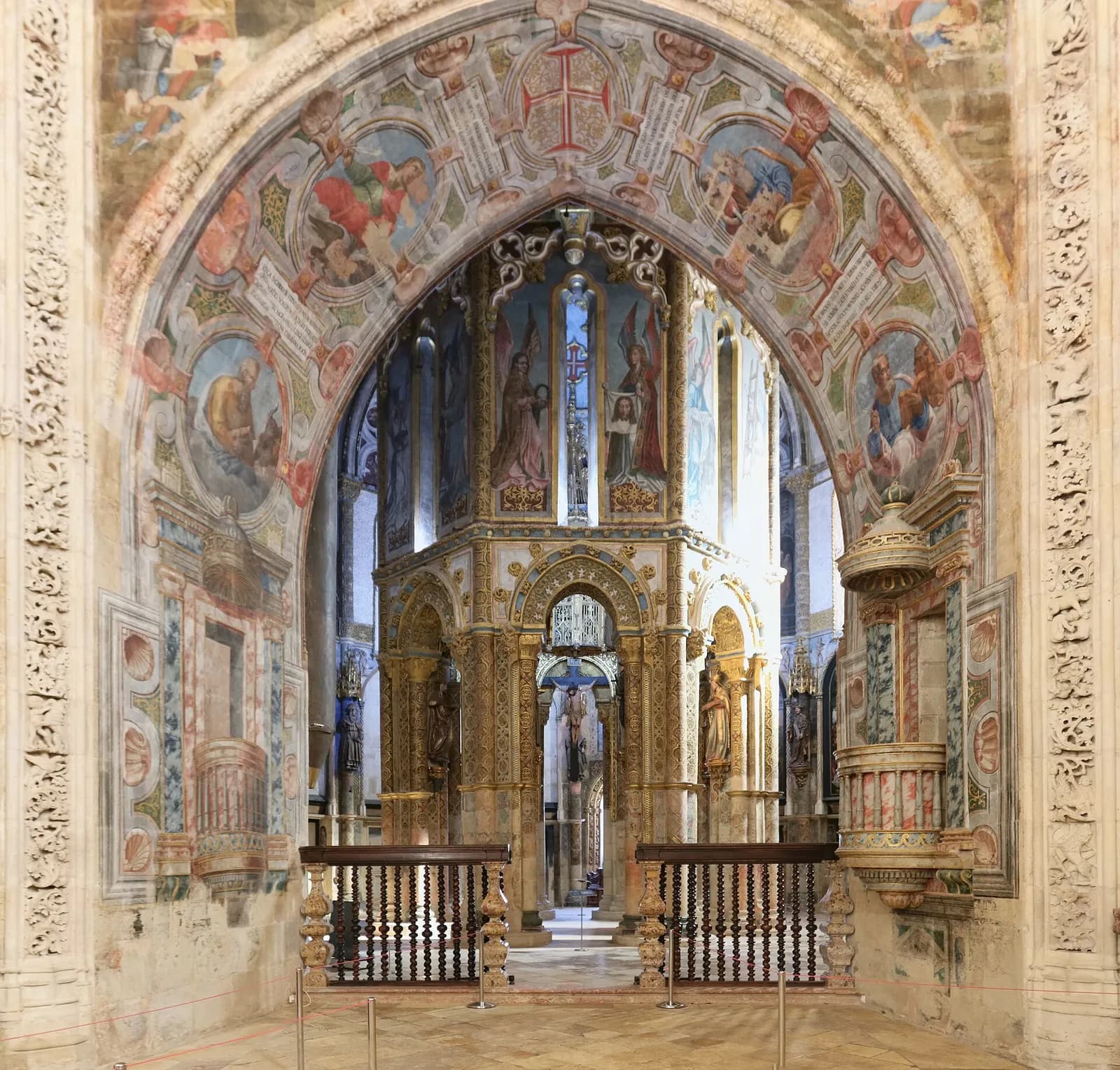 Looking through a tall arch covered in painted saints and patterns into a round church. At its centre stands a gilded, many-sided structure with painted panels and statues, lit by high windows.