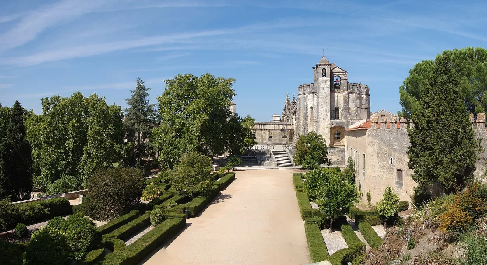 A wide gravel avenue between clipped box hedges and tall trees leads up to a stone complex, where a tall many-sided church with a bell turret rises above long convent walls under a blue sky.