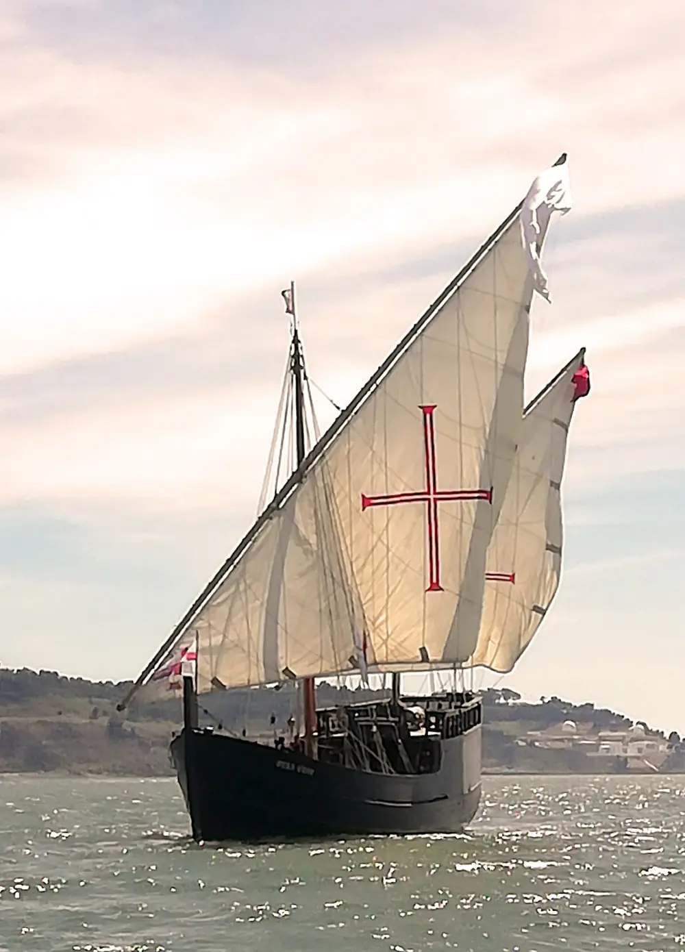 A small wooden sailing ship with two triangular sails, the larger one painted with a red cross, sails across a wide grey river with low hills behind.