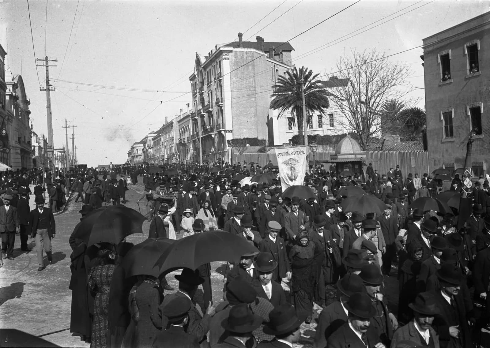 A large crowd of men in bowler hats and women in shawls walks along a city street under umbrellas, with tram wires overhead and a palm tree behind a fence. A banner with a painted figure and a flag are carried in the middle of the procession.