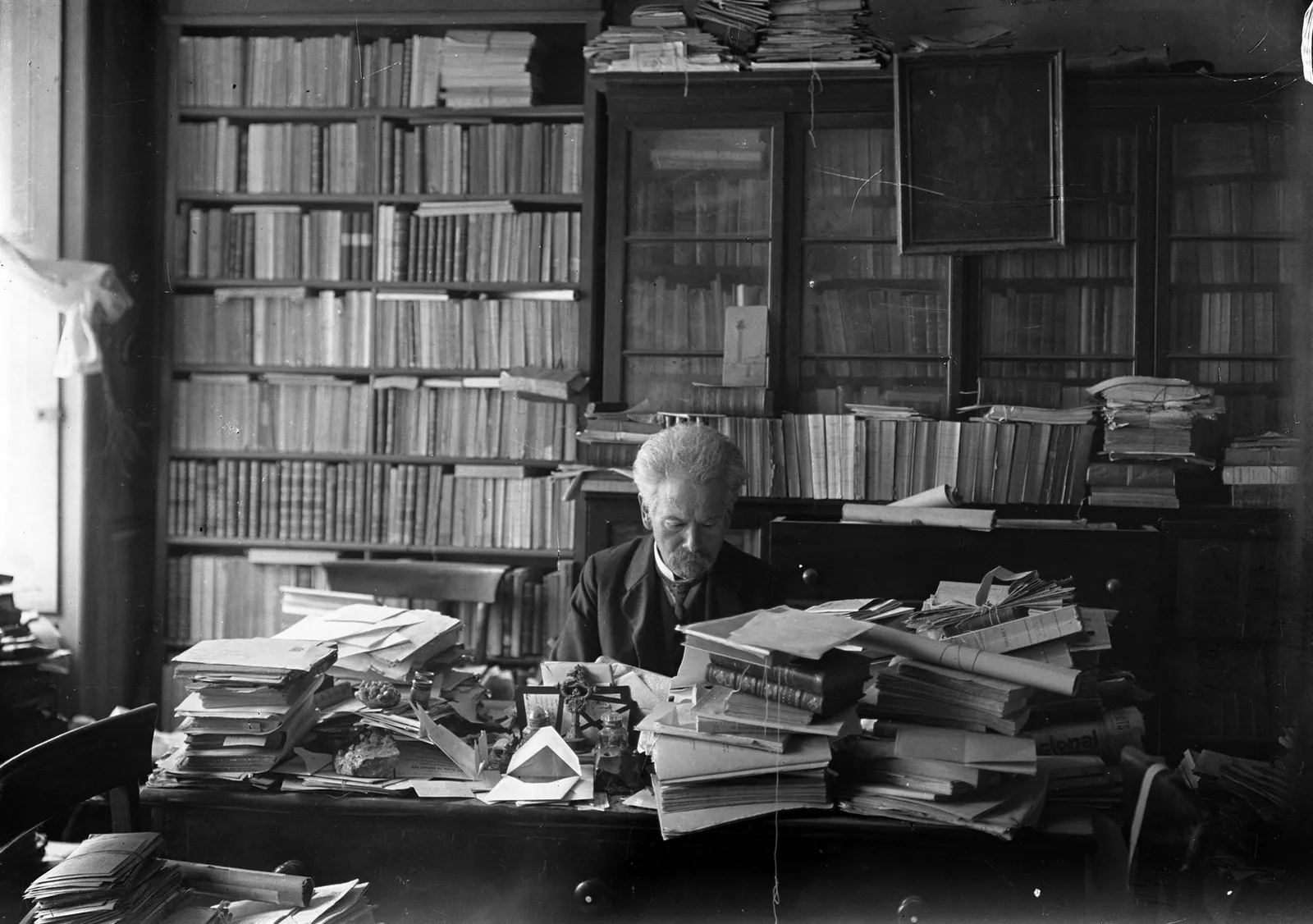 An elderly man with a shock of white hair and a white beard sits reading behind a desk piled high with books, letters and bundles of papers tied with string. Shelves and glass-fronted bookcases crammed with books fill the wall behind him.