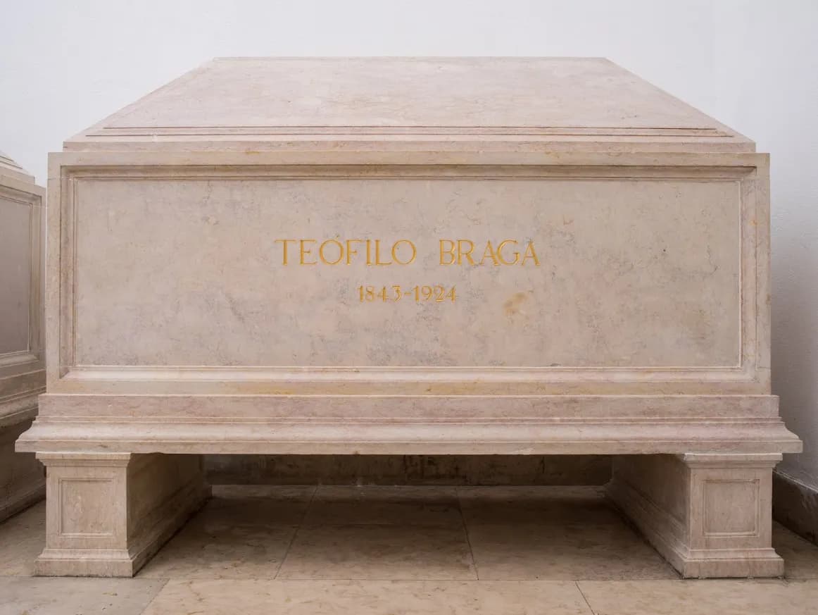 A plain rectangular tomb of pale pink marble on two square feet stands against a white wall. Gold capital letters on its side read TEÓFILO BRAGA, with the dates 1843-1924 beneath.