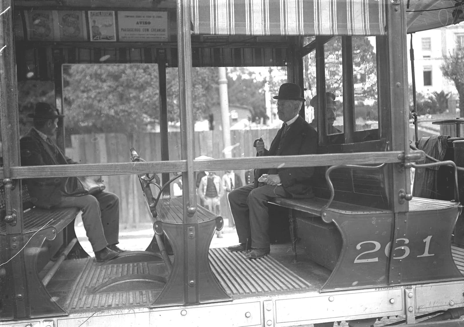 An elderly man with a white beard, in a bowler hat and dark coat, sits on a slatted wooden bench of an open tramcar, holding a walking stick and some papers. The number 261 is painted on the side of the car. Another passenger sits facing him and a few onlookers stand in the street behind.