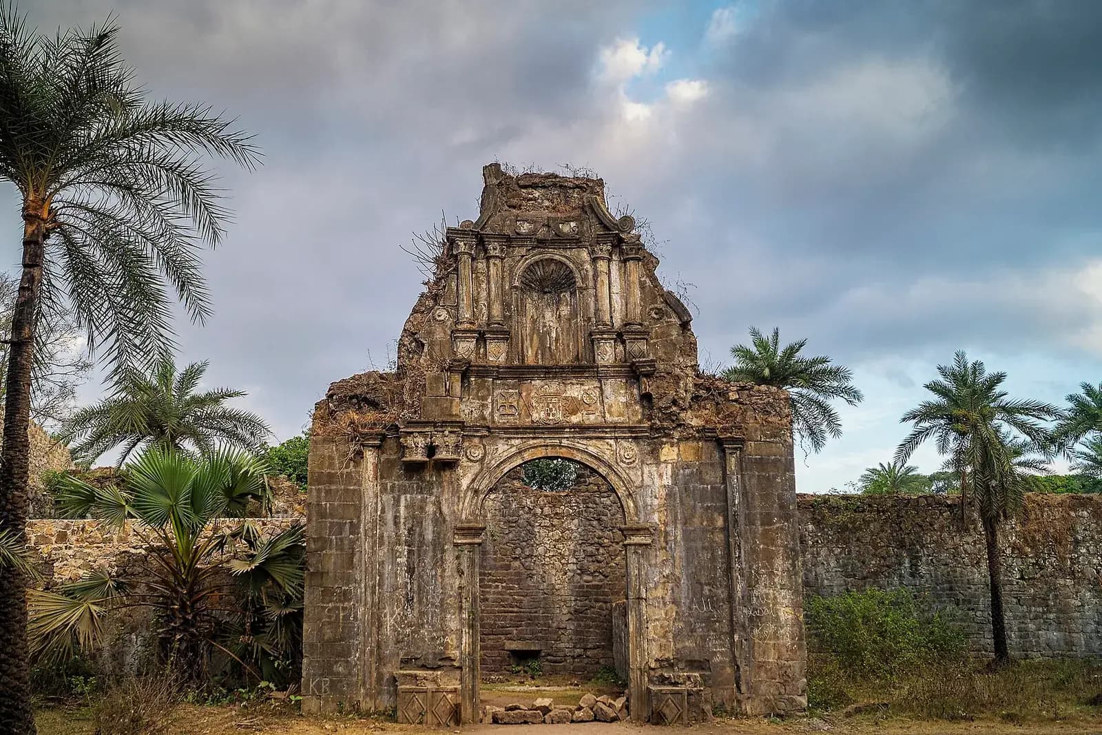 A tall, crumbling stone gateway with a round arch and a carved upper storey stands among palm trees, with a stretch of rough stone wall to the right and a cloudy sky behind.
