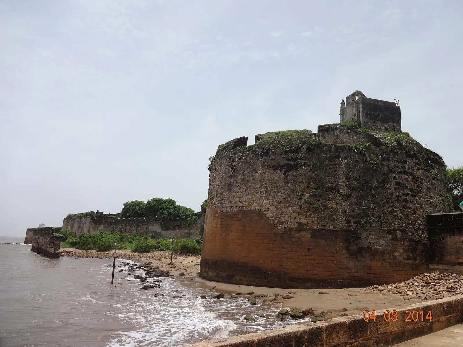 A massive round stone bastion, its lower courses stained brown by the sea, rises from a rocky beach, with a small square watchtower on top and a long line of walls running off to the left along the shore.