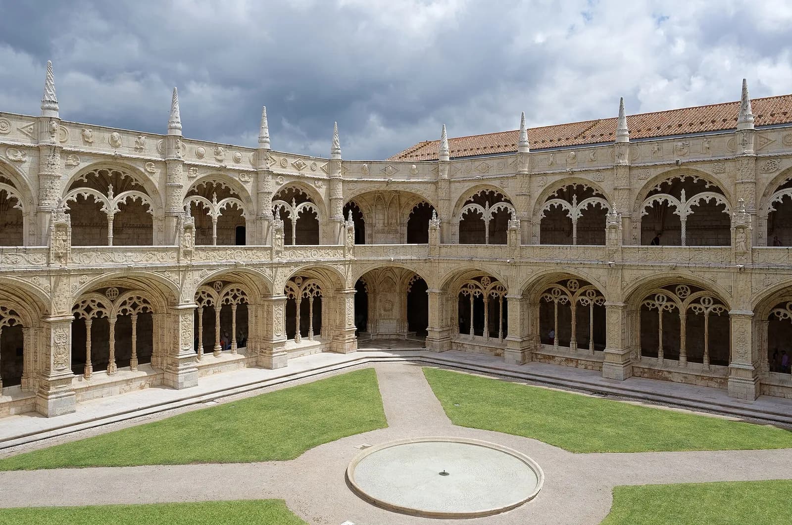 A square courtyard of pale limestone surrounded by two storeys of richly carved arcades, with a lawn crossed by paths and a round basin in the centre, under a cloudy sky.