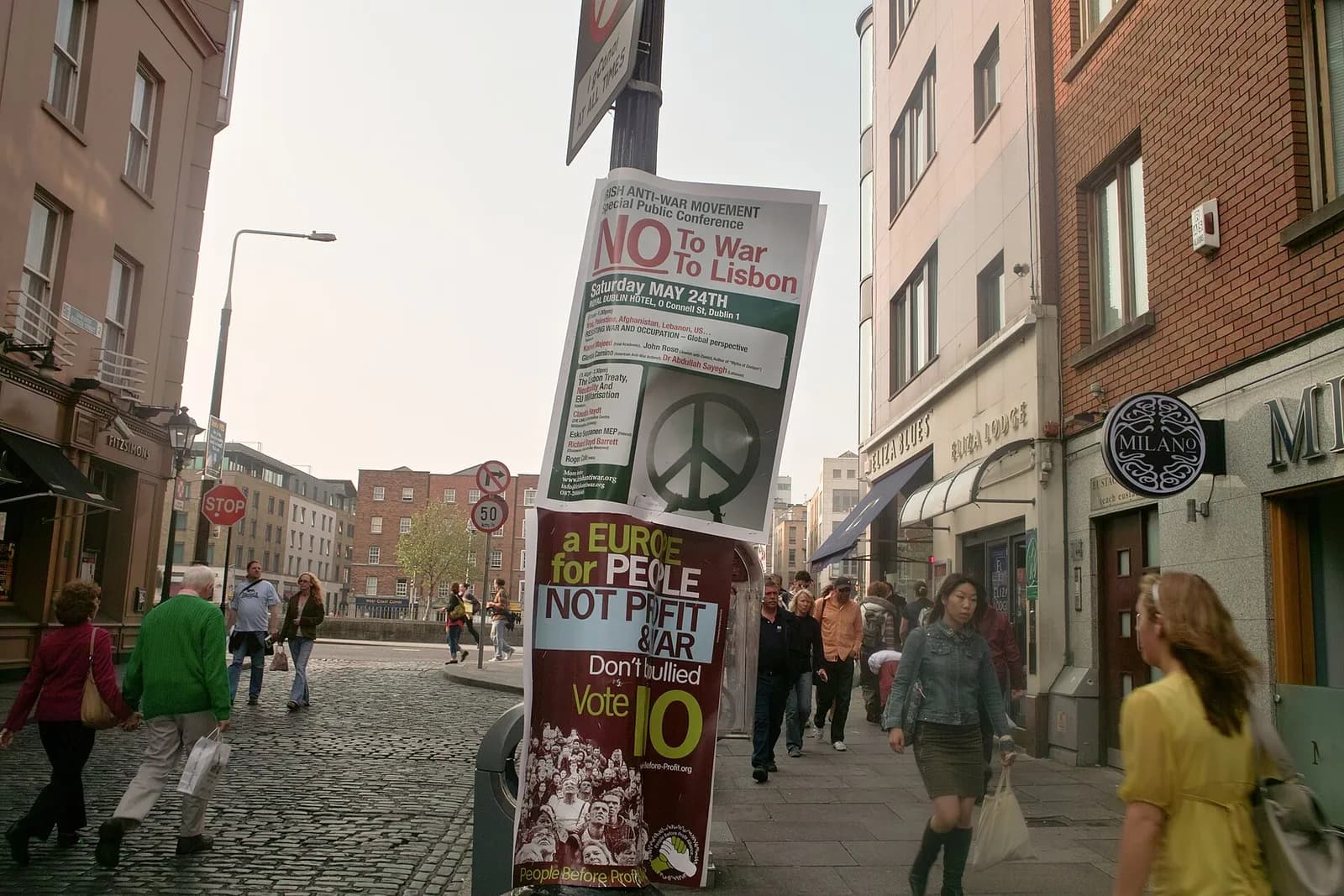 Two posters tied to a lamp post on a cobbled Dublin street. The upper one, from the Irish Anti-War Movement, reads “No to war, no to Lisbon”; the lower one reads “A Europe for people, not profit and war. Don’t be bullied. Vote no”. Shoppers walk past.