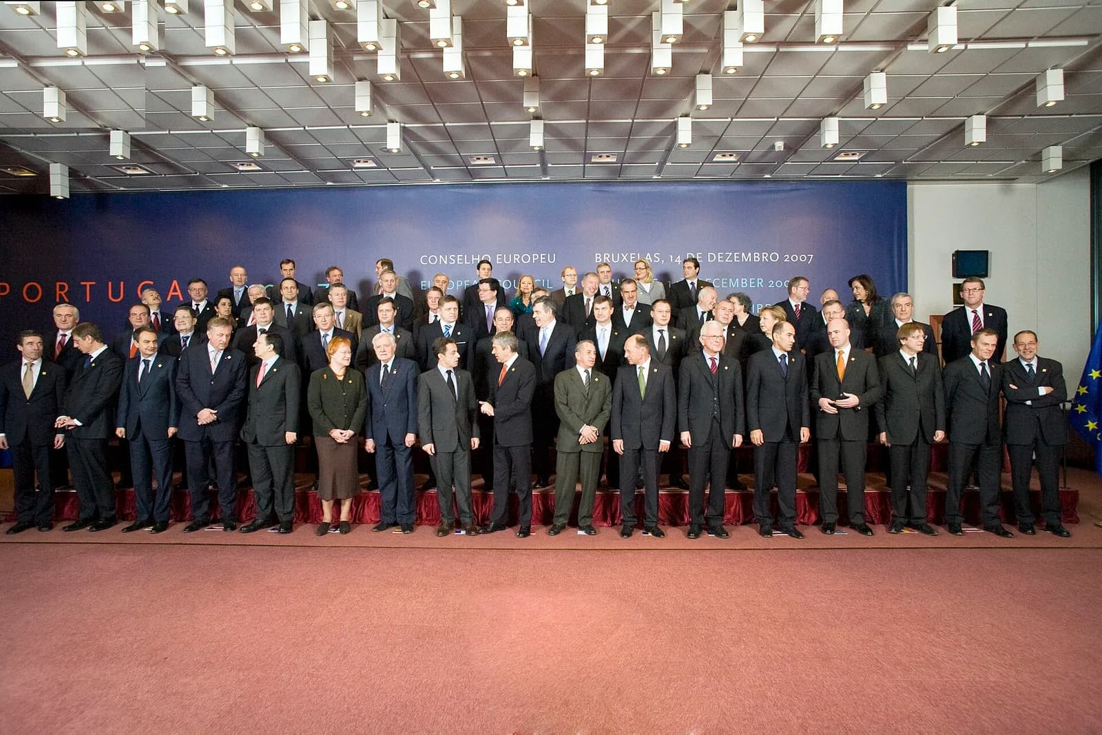 About sixty people, nearly all men in dark suits, stand in four rows on tiered steps in front of a blue backdrop reading “Conselho Europeu, Bruxelas, 14 de Dezembro 2007”, with “Portugal” on the left.