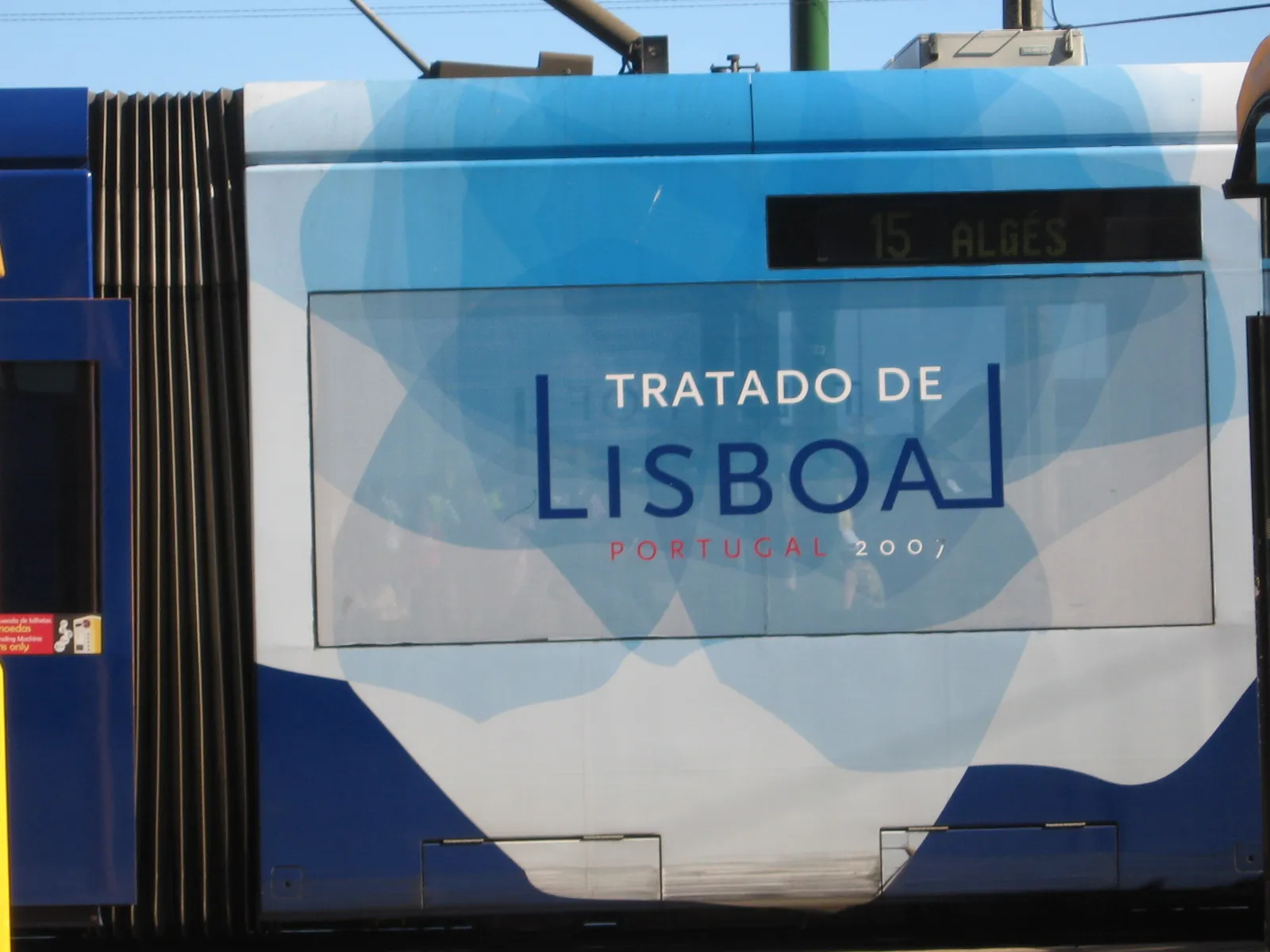 The side of a blue and white articulated tram with a large panel reading “Tratado de Lisboa, Portugal 2007”; the destination display above it reads “15 Algés”.