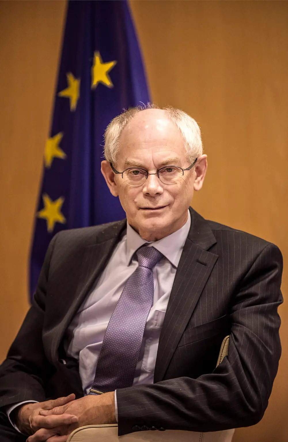 A grey-haired man in glasses and a dark pinstriped suit with a purple tie sits in an armchair in front of a blue EU flag with yellow stars and a wood-panelled wall.