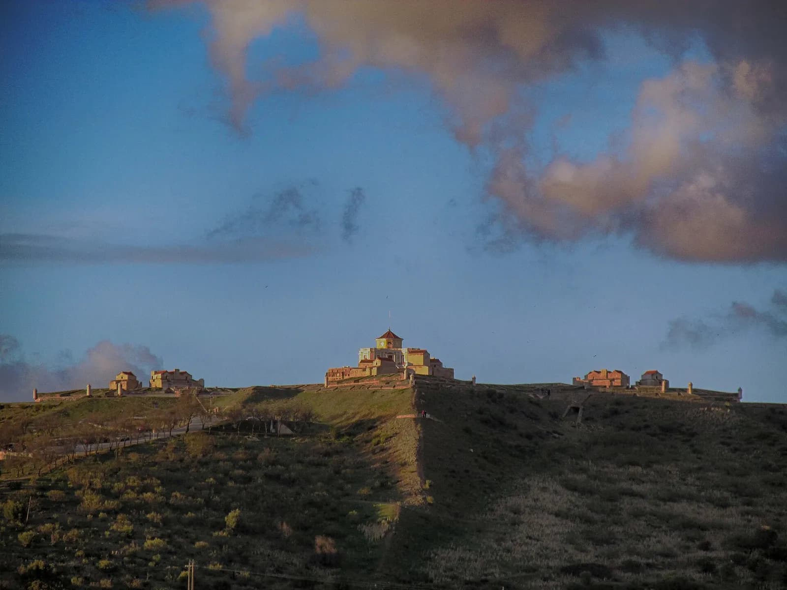 A low fortress of ochre buildings, with a central tower under a pointed roof, sits on the crest of a grassy hill in late-afternoon light, under a blue sky with pink clouds.