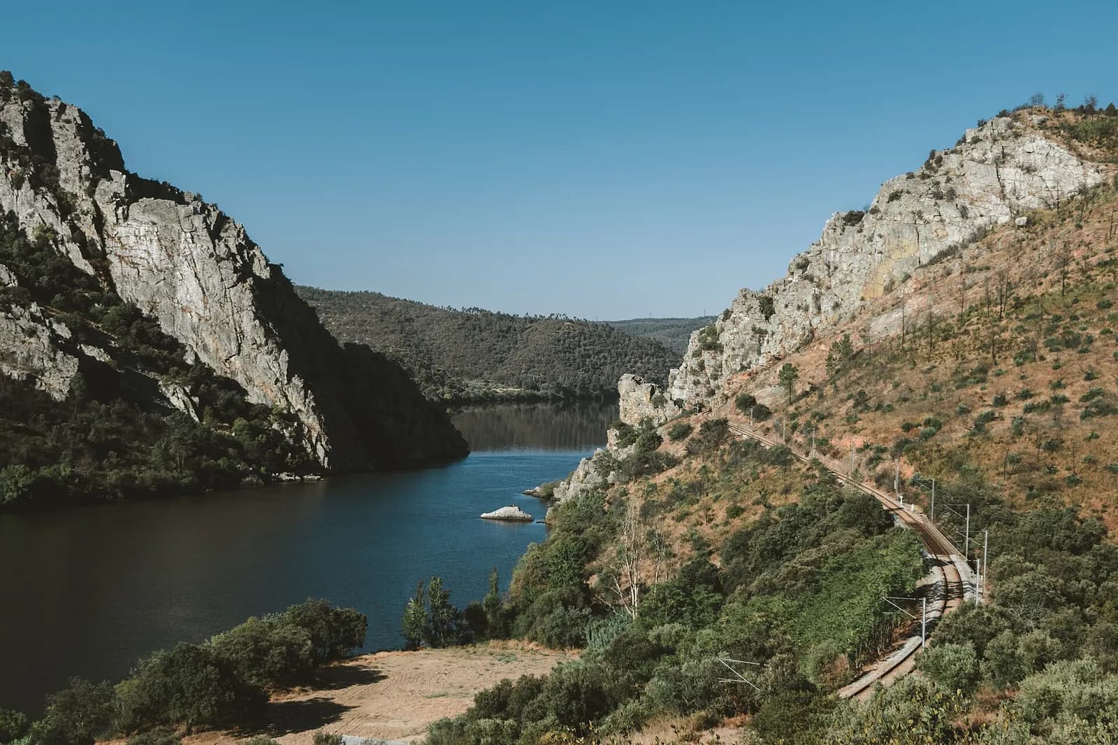 A wide river runs between two steep rocky hills covered in scrub, under a clear blue sky. A railway line curves along the slope on the right.