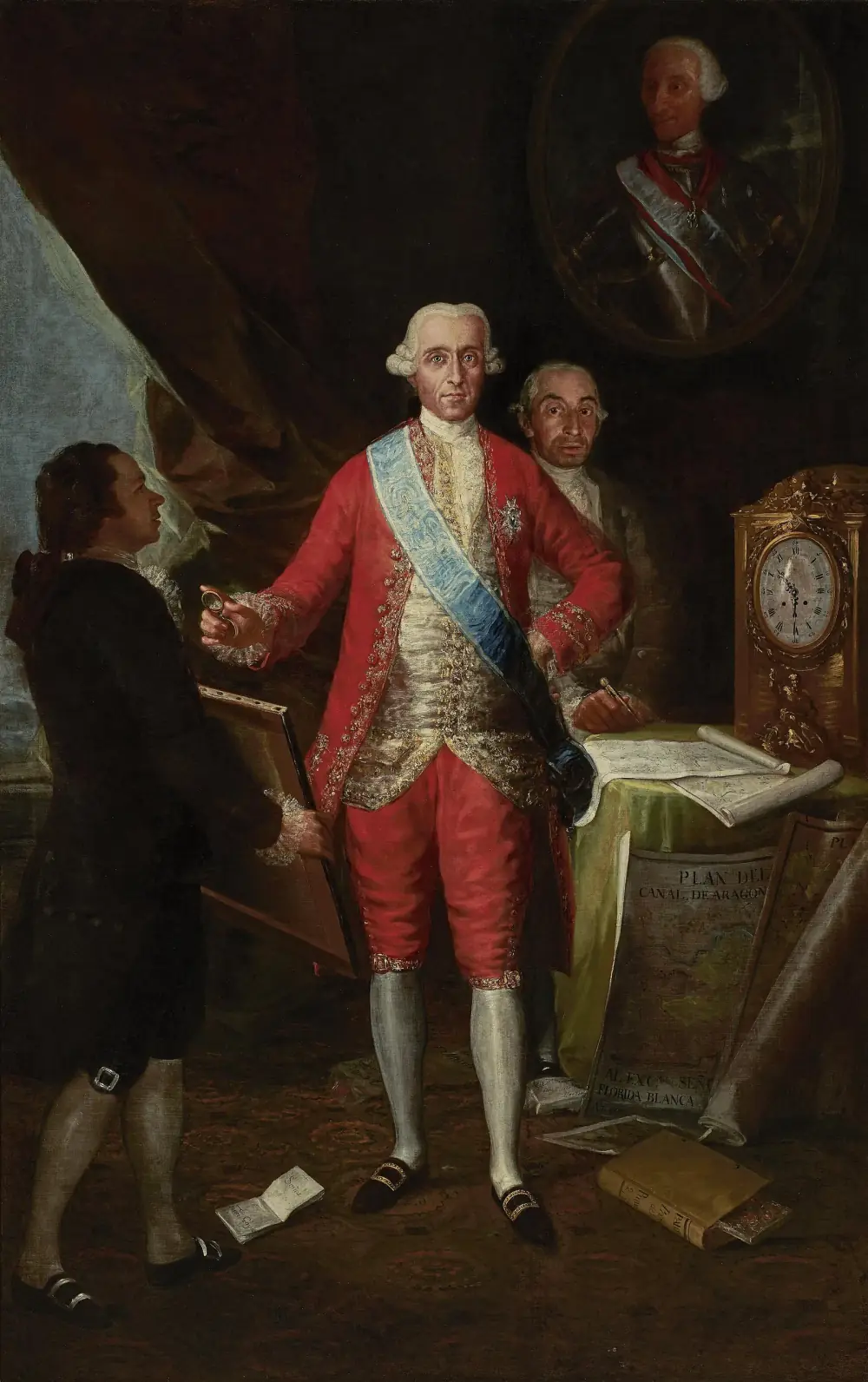 Full-length painting of a man in a red embroidered court suit and a pale blue sash, standing in a dark room beside a table covered with maps and a gilt clock. A second man stands behind him and a third, holding a framed picture, faces him from the left. An oval portrait of the king hangs on the wall above, and a large plan leaning against the table is titled Plan del Canal de Aragón.