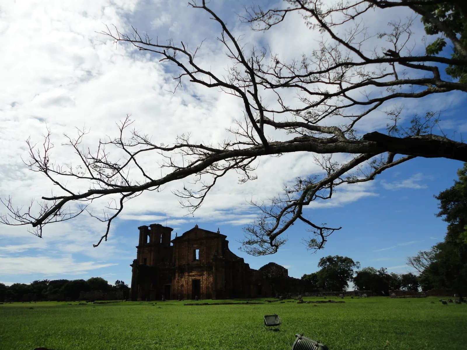 The roofless stone façade and bell tower of a large ruined church stand at the far end of a wide, flat lawn under a blue sky with white clouds. Bare tree branches stretch across the top of the picture.