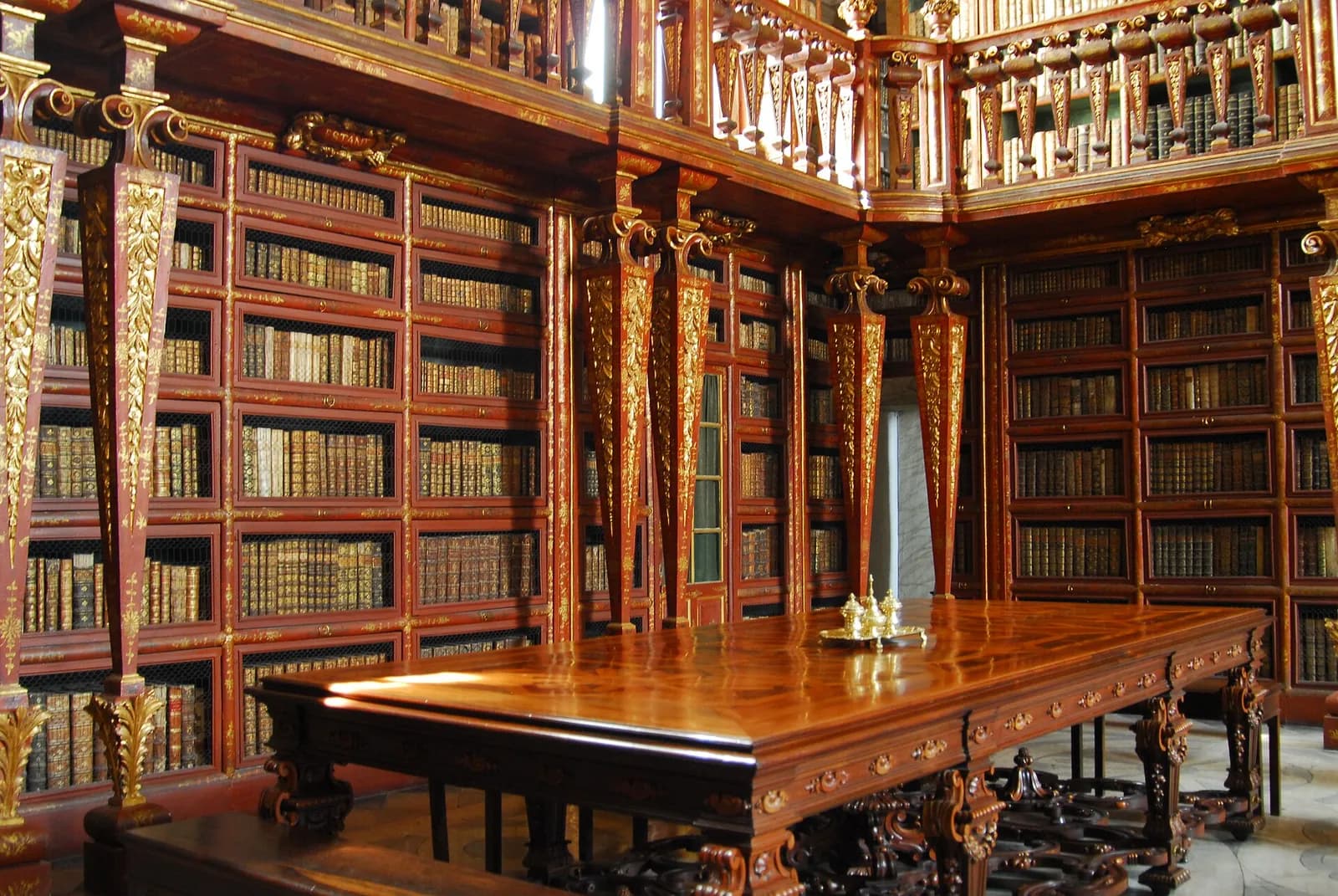 A library interior of red and gold lacquered bookcases on two levels, with gilded pillars and a balustraded gallery above. Rows of old leather-bound books fill the shelves, and a large polished wooden table with a small gilt object stands in the foreground.