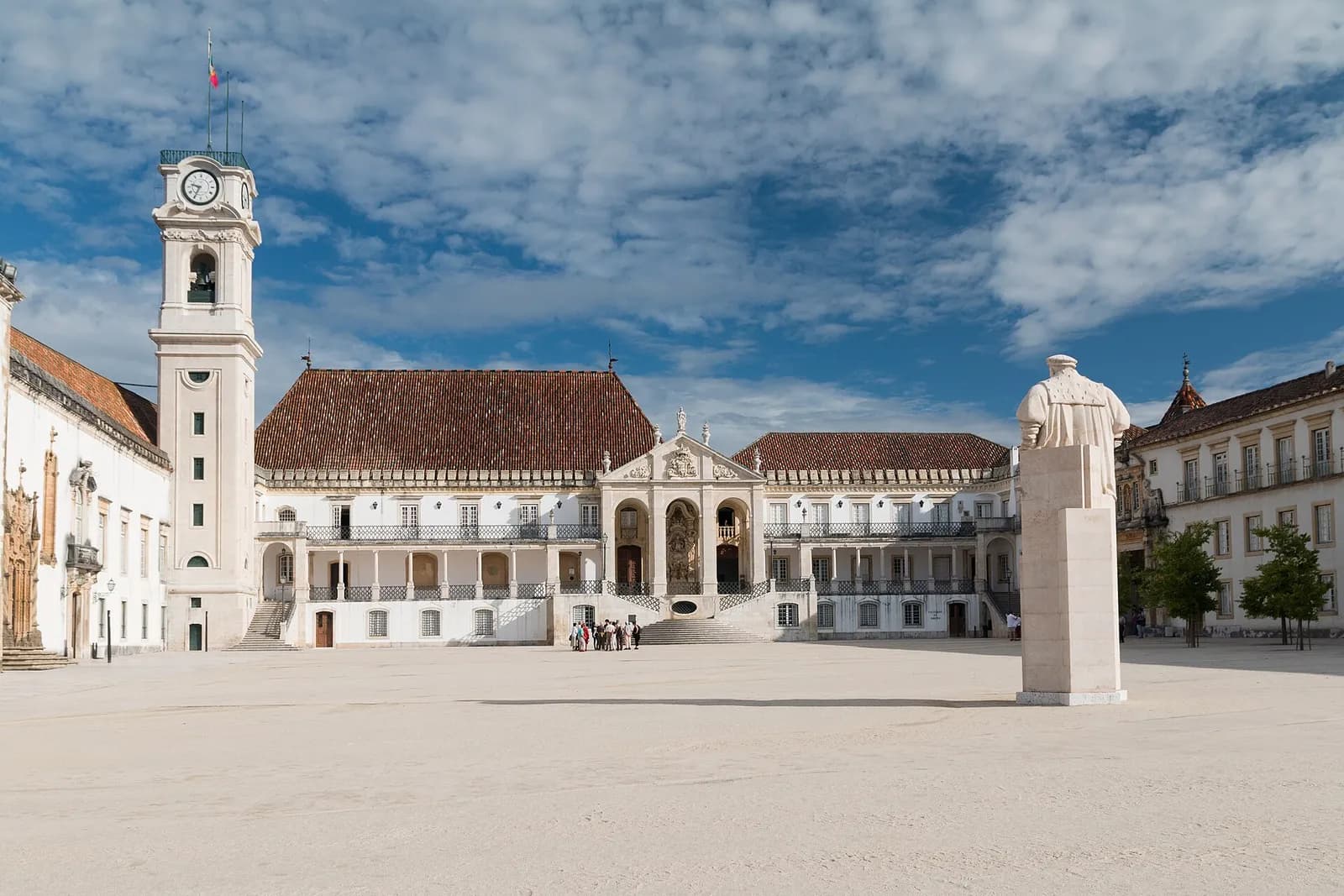 A wide, pale stone courtyard under a blue sky with white clouds. On the left a white clock tower; across the back a long white palace with a red tiled roof, an open gallery of columns and a central staircase; on the right a white statue of a man in a cloak on a tall plinth.