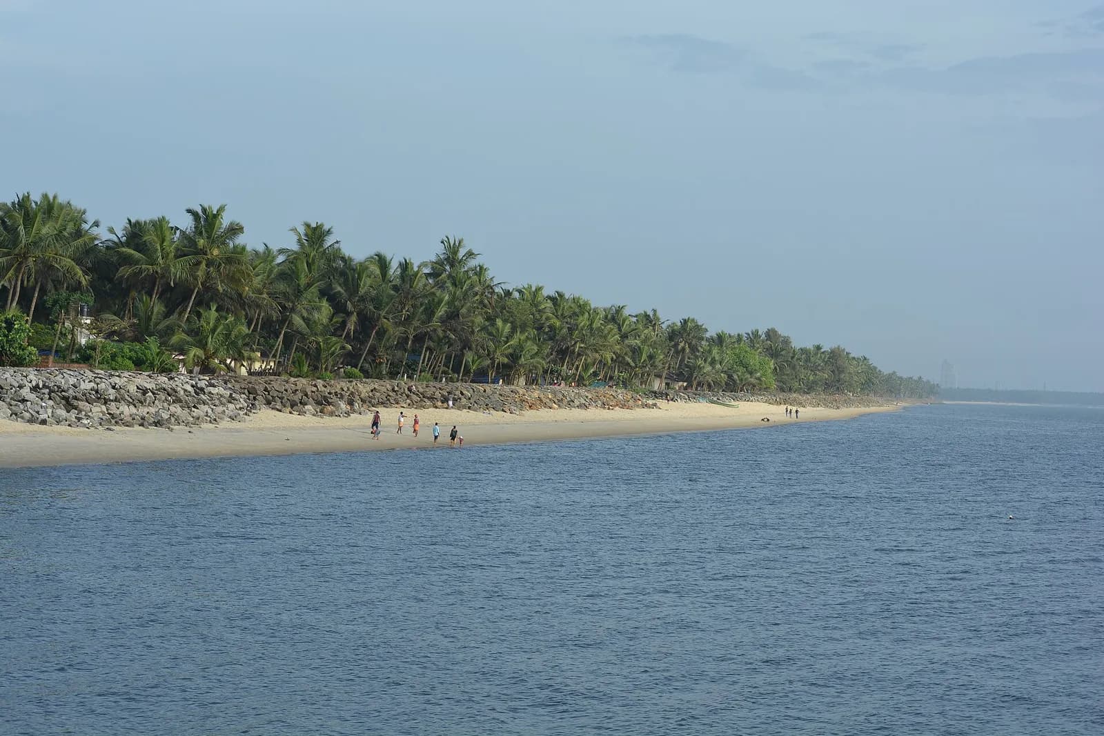 A long sandy beach lined with a rock sea wall and dense coconut palms curves away into haze, with a few people walking at the water's edge and a calm grey-blue sea in the foreground.