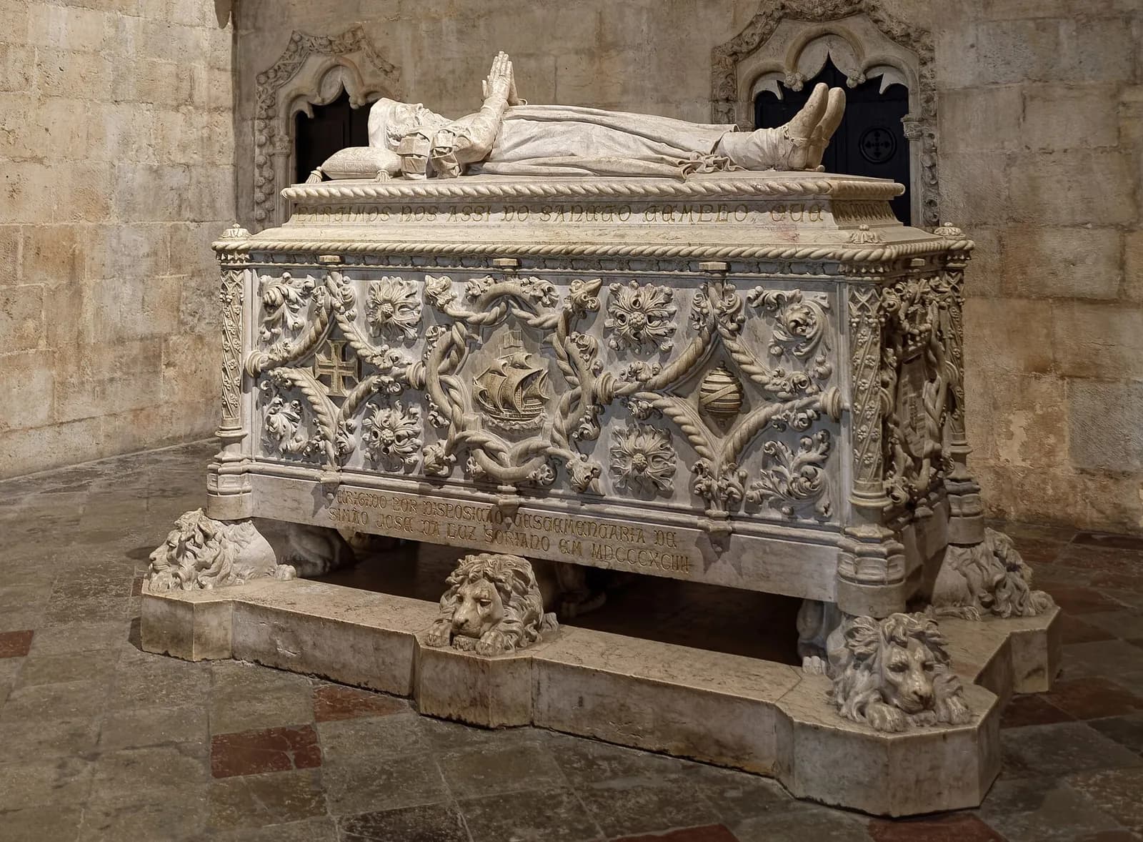A carved white stone tomb chest resting on crouching lions, decorated with twisted ropes, foliage, a sailing ship, a cross and an armillary globe, with a recumbent figure in prayer lying on the lid.