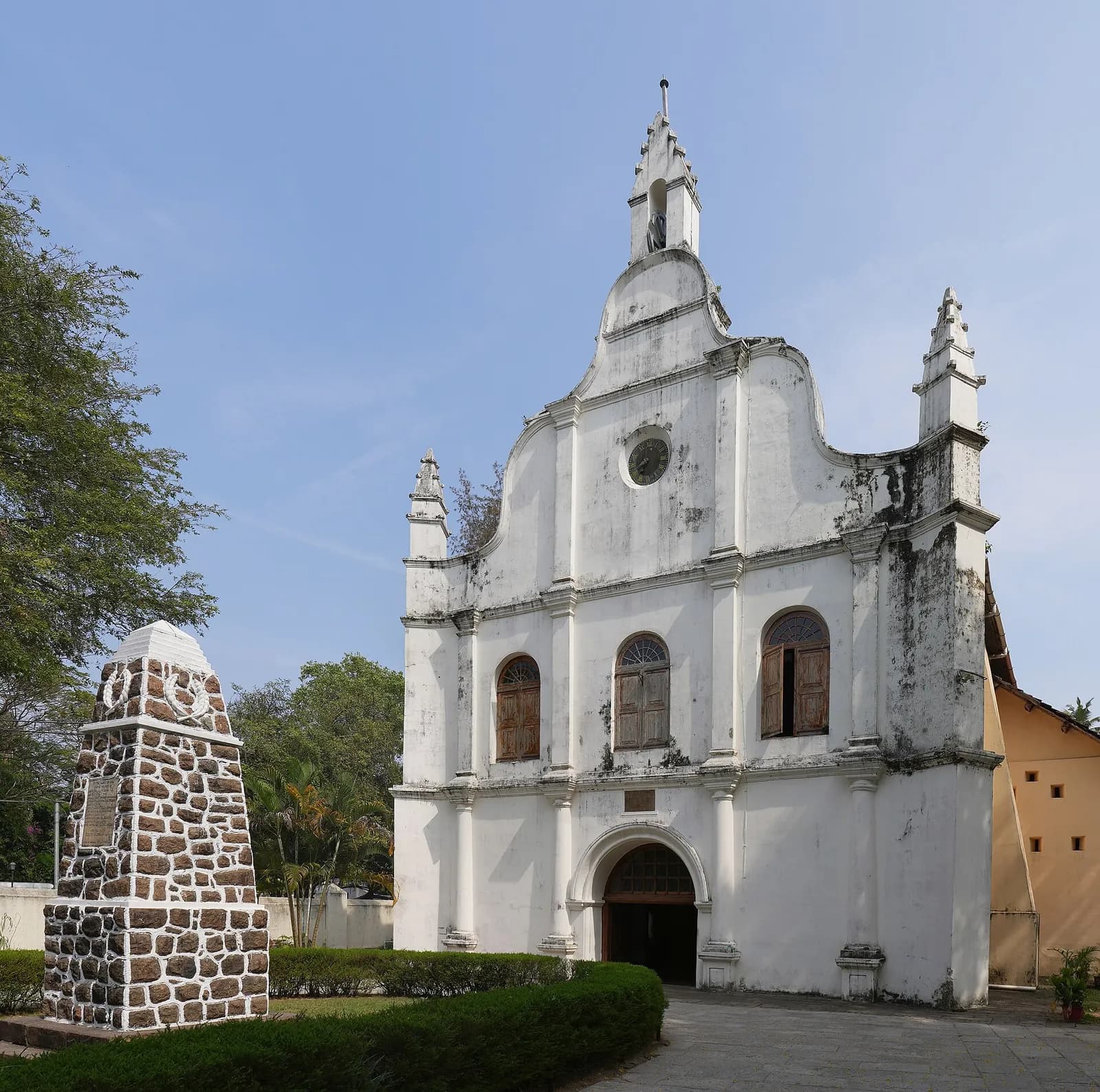 The whitewashed gabled facade of a small church with arched windows and a bell turret against a blue sky, with a stone memorial pillar on the left.
