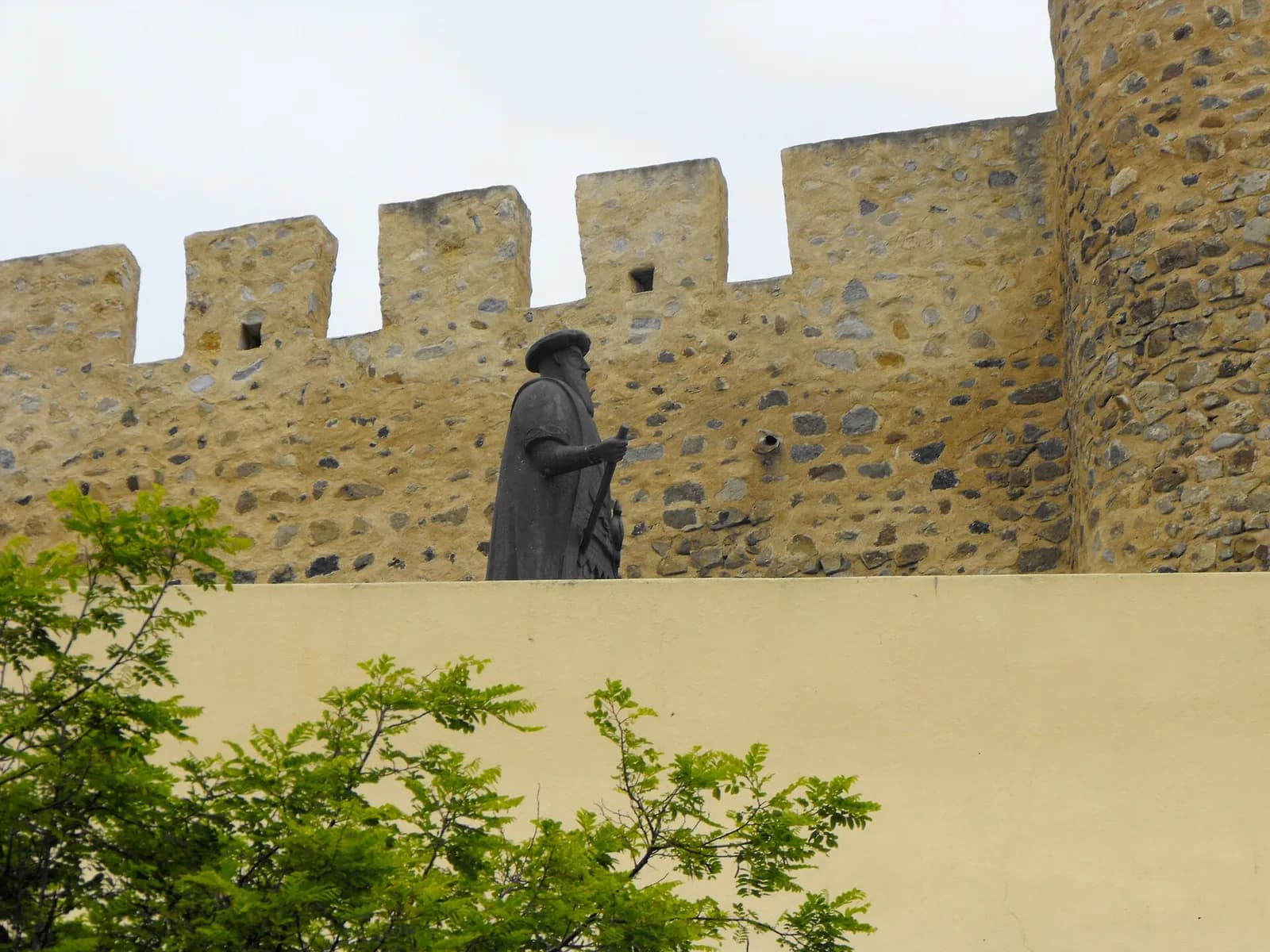 A dark statue of a bearded man in a cloak and flat cap, holding a sword hilt, stands on a terrace in front of crenellated stone castle walls, with green leaves in the foreground.