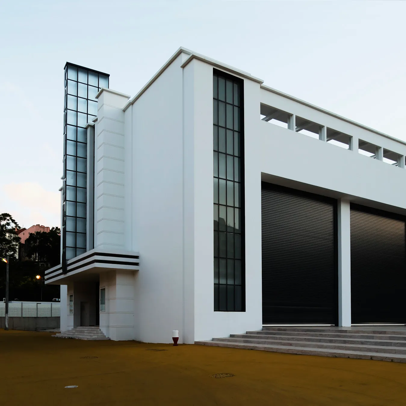 A white modernist building with a tall glass tower at one corner, a tall narrow window, an open gallery along the top and two large dark roller doors at the top of a flight of steps, seen across an empty yard at dusk.