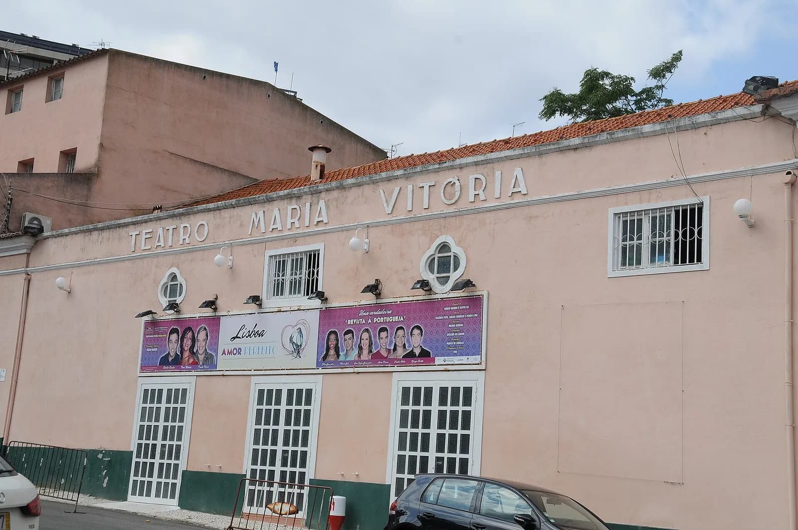 The long, low pink facade of a theatre with the words Teatro Maria Vitória in raised letters along the top. Above its white-framed doors hangs a row of banners with smiling performers advertising a revista à portuguesa.