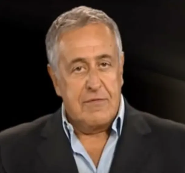 Head and shoulders of a grey-haired man with dark eyebrows in a dark jacket and pale blue open-necked shirt, against a black background.