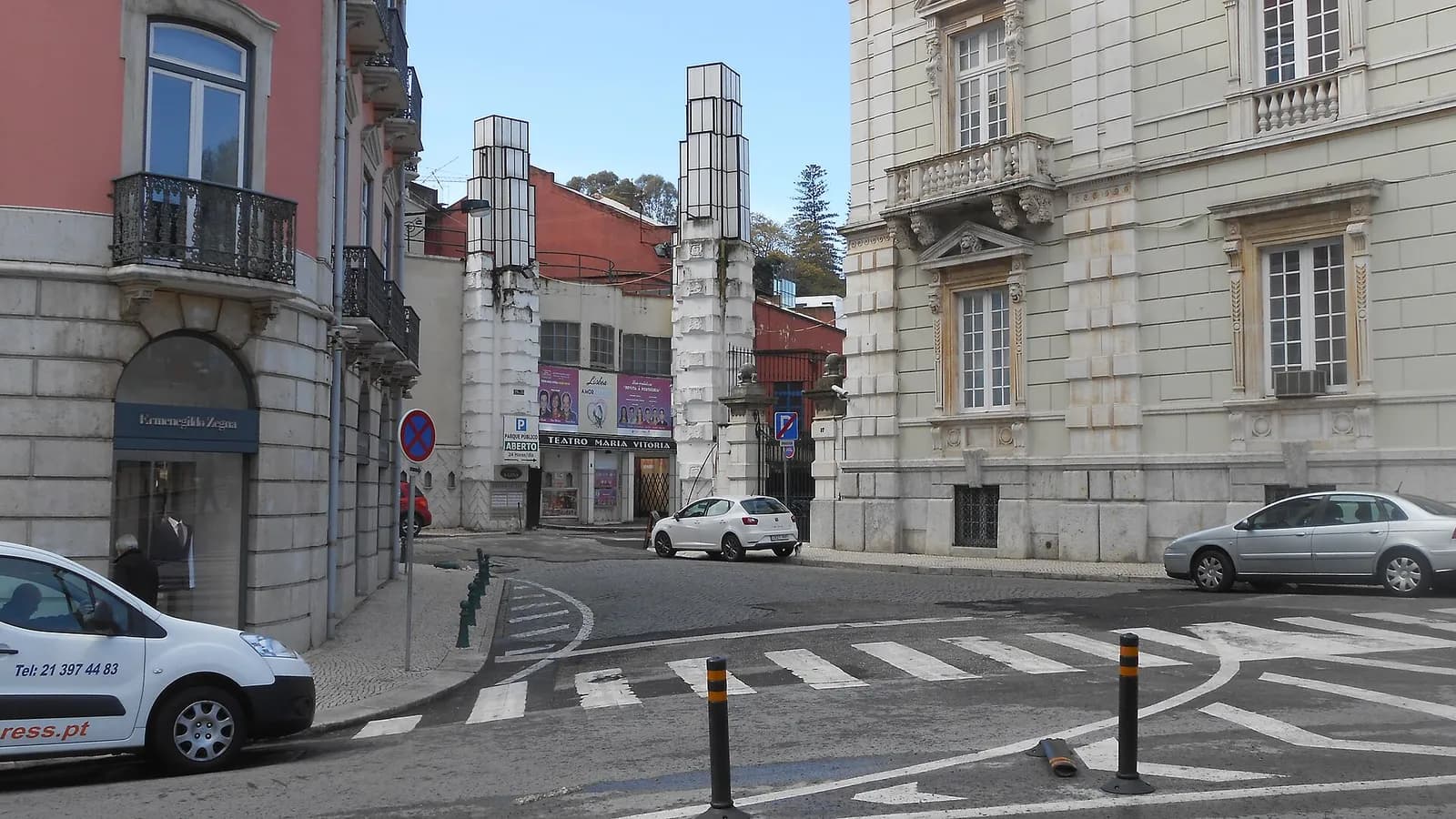 A short street runs between a pink building and a grand pale stone mansion towards two tall white pylons topped with glass lanterns. Between them is a gateway, and at the end of the drive a theatre sign reads Teatro Maria Vitória.