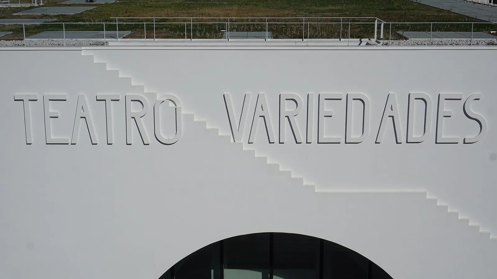 A smooth white wall with the words Teatro Variedades cut into it in tall thin capitals, a stepped diagonal line running across it and the top of a round-arched glass doorway below. A planted roof terrace with railings is visible above.