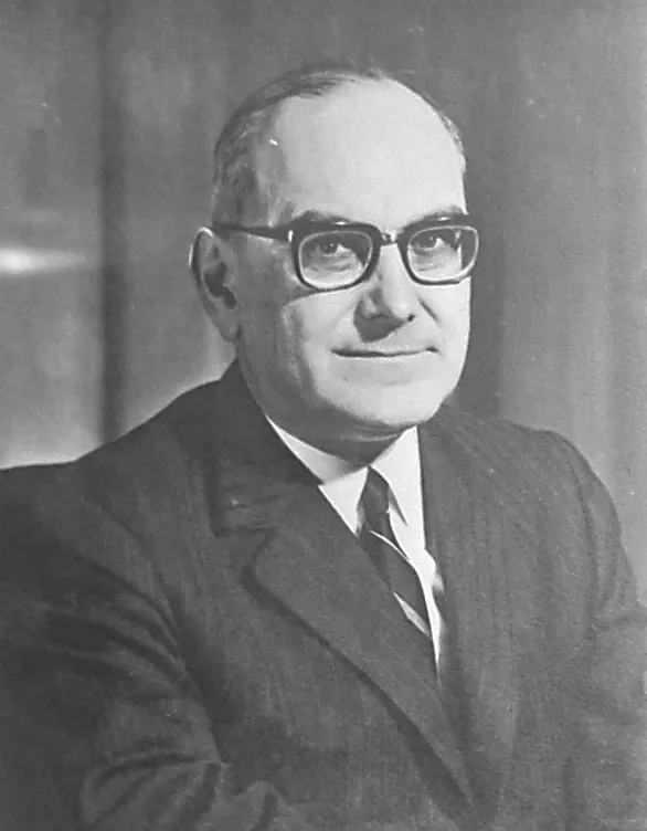 Black-and-white portrait of a balding man in thick dark-rimmed glasses, a dark suit and striped tie, smiling slightly.