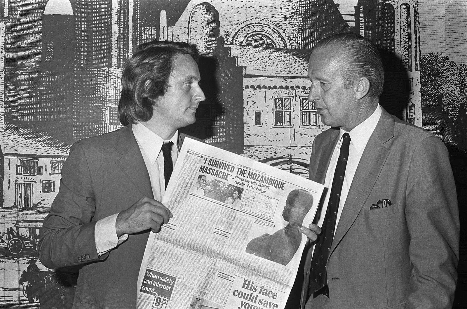 Two men in suits and ties stand in front of a mural of old buildings. The younger man, with long hair, holds open a newspaper page headlined I survived the Mozambique massacre, with a photograph of a boy.