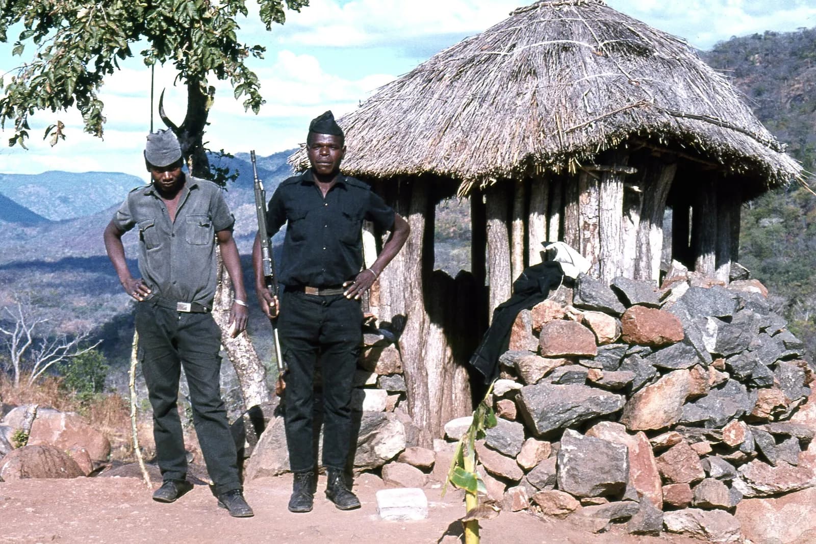 Two African men in caps stand on dusty ground beside a thatched wooden hut and a pile of stones, one in a grey shirt with his hands on his hips, the other in a black uniform holding a rifle upright. Wooded hills rise behind them.