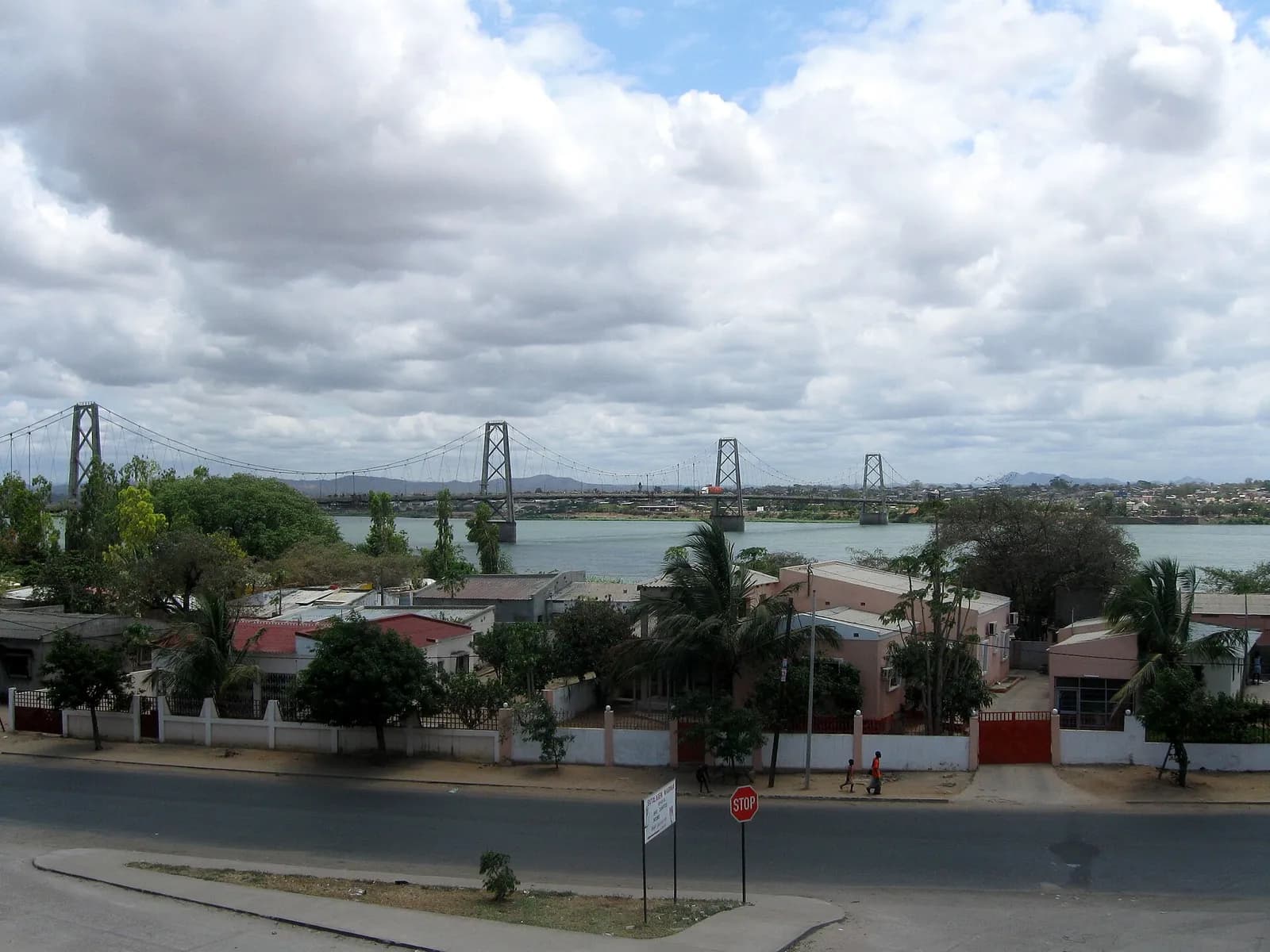 A long suspension bridge with four pylons crosses a wide river under a sky of heavy white clouds. In the foreground are a road with a stop sign and low houses with pink walls among palm trees.