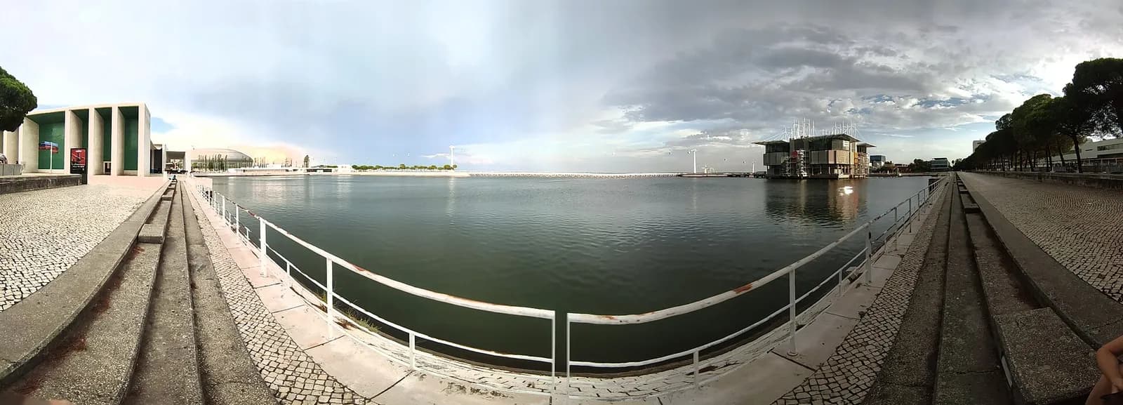 Wide panoramic view of a calm rectangular dock framed by stone quays, with a low modern pavilion on the left and a building standing in the water on the right under a cloudy sky.