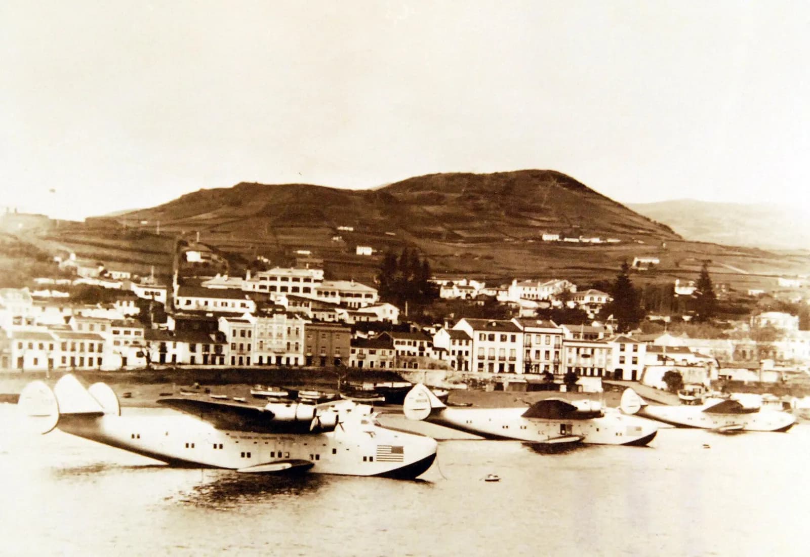 Three large flying boats with triple tail fins moored in a harbour in front of a waterfront town of white houses, with a hill behind. The photograph is sepia-toned.