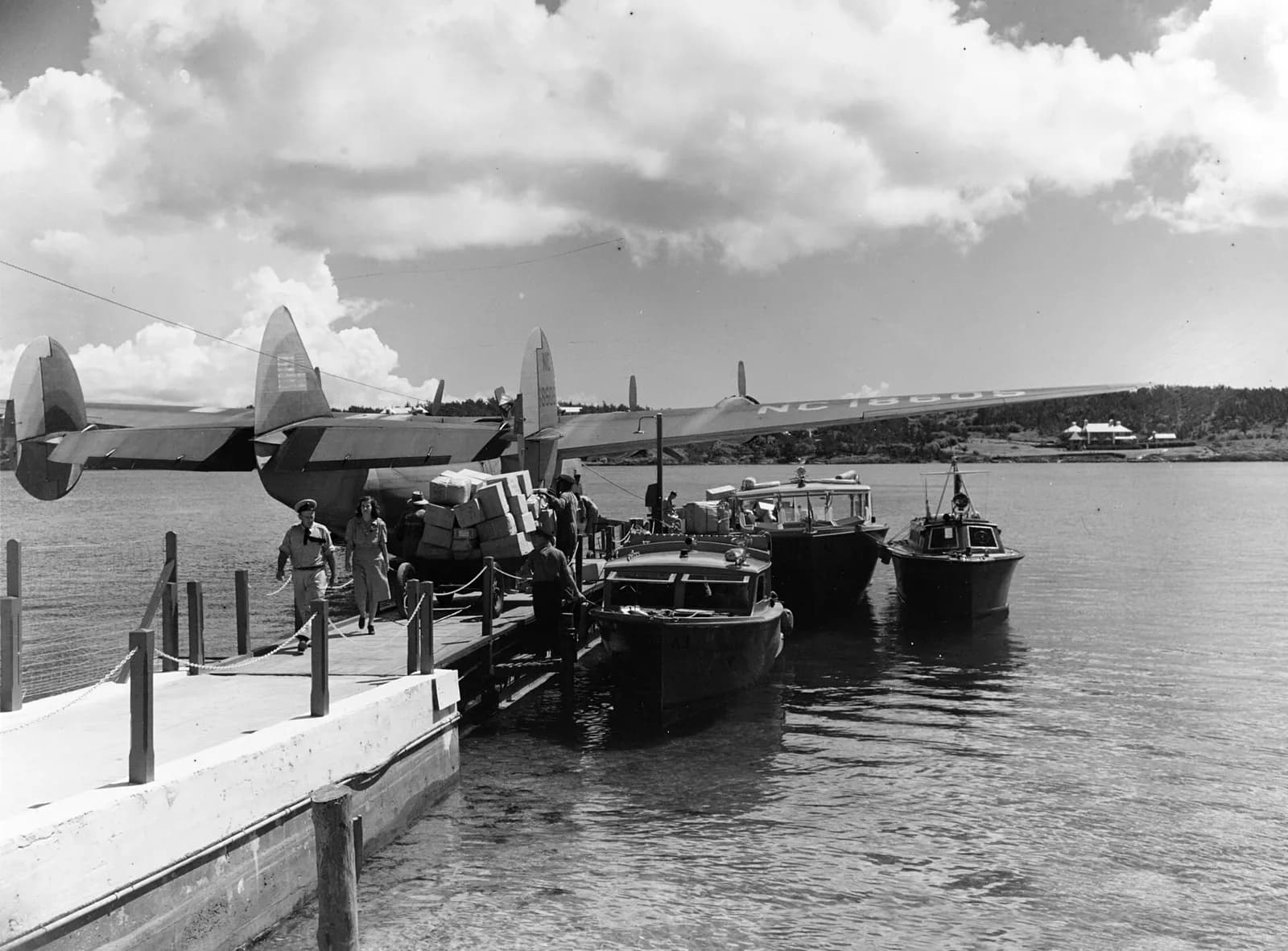 A flying boat with a camouflaged upper surface and NC 18605 on its wing moored beside a small pier. Men load boxes on a trolley while a man and a woman walk along the jetty past three motor launches.