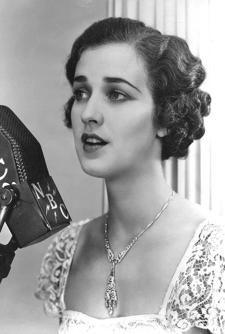 Black-and-white studio portrait of a young woman with short dark waved hair, a lace dress and a long pendant necklace, singing into an NBC microphone.