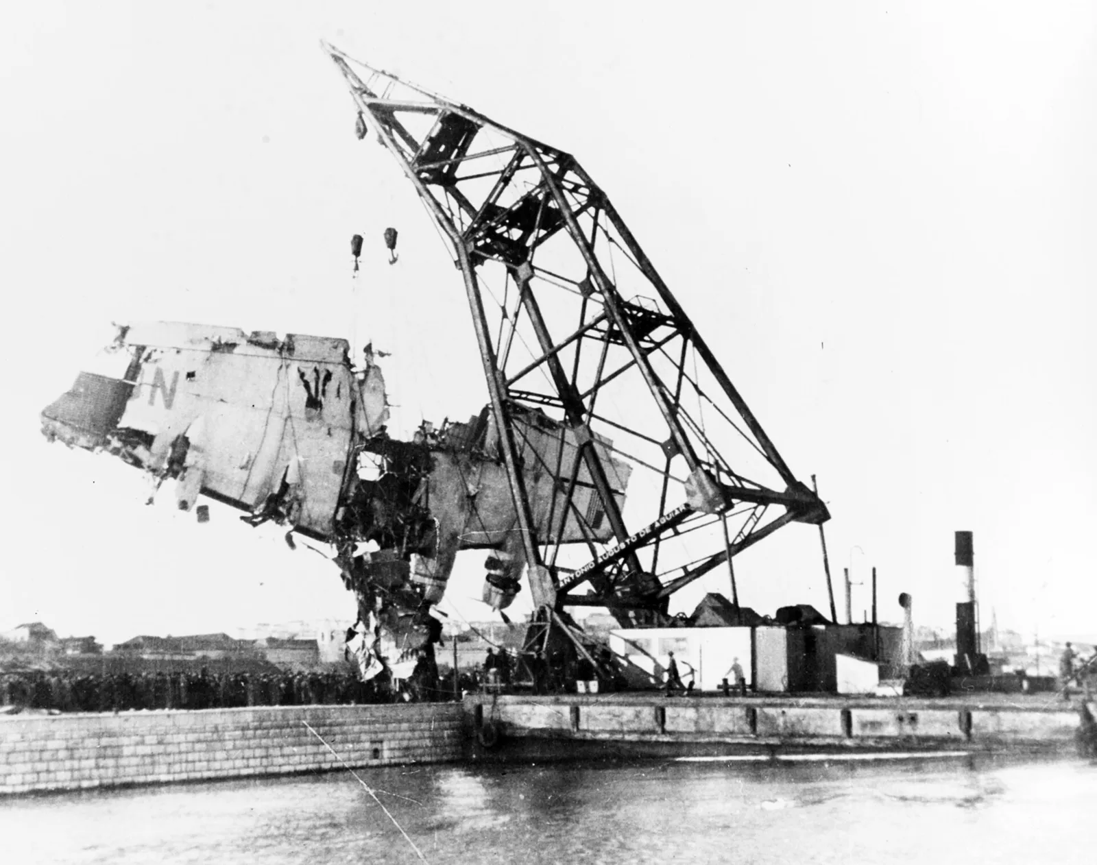 A tall lattice floating crane lifts a broken section of an aircraft hull out of the water beside a stone quay lined with a dense crowd of onlookers.
