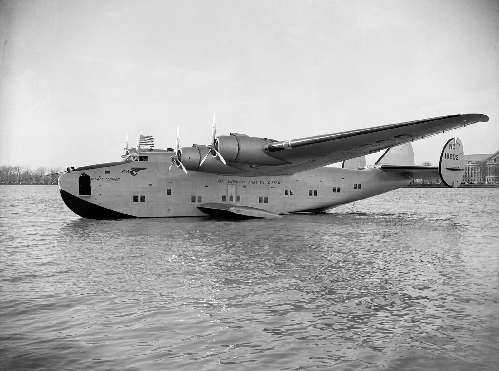 A large four-engined flying boat floats on calm water, moored by a line at the bow. Yankee Clipper and Pan American Airways System are painted on the hull and NC 18603 on the tail fin.