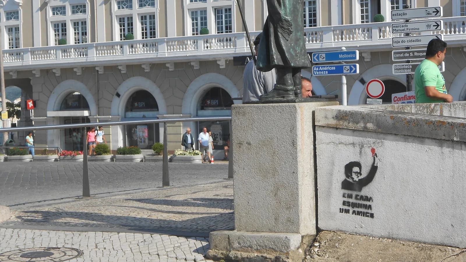 A black stencil on a low concrete wall in a cobbled square: a man with curly hair and glasses raises his arm holding a red flower. Under him are the words Em cada esquina um amigo. Behind are a bronze statue's base, road signs and an arcaded building.