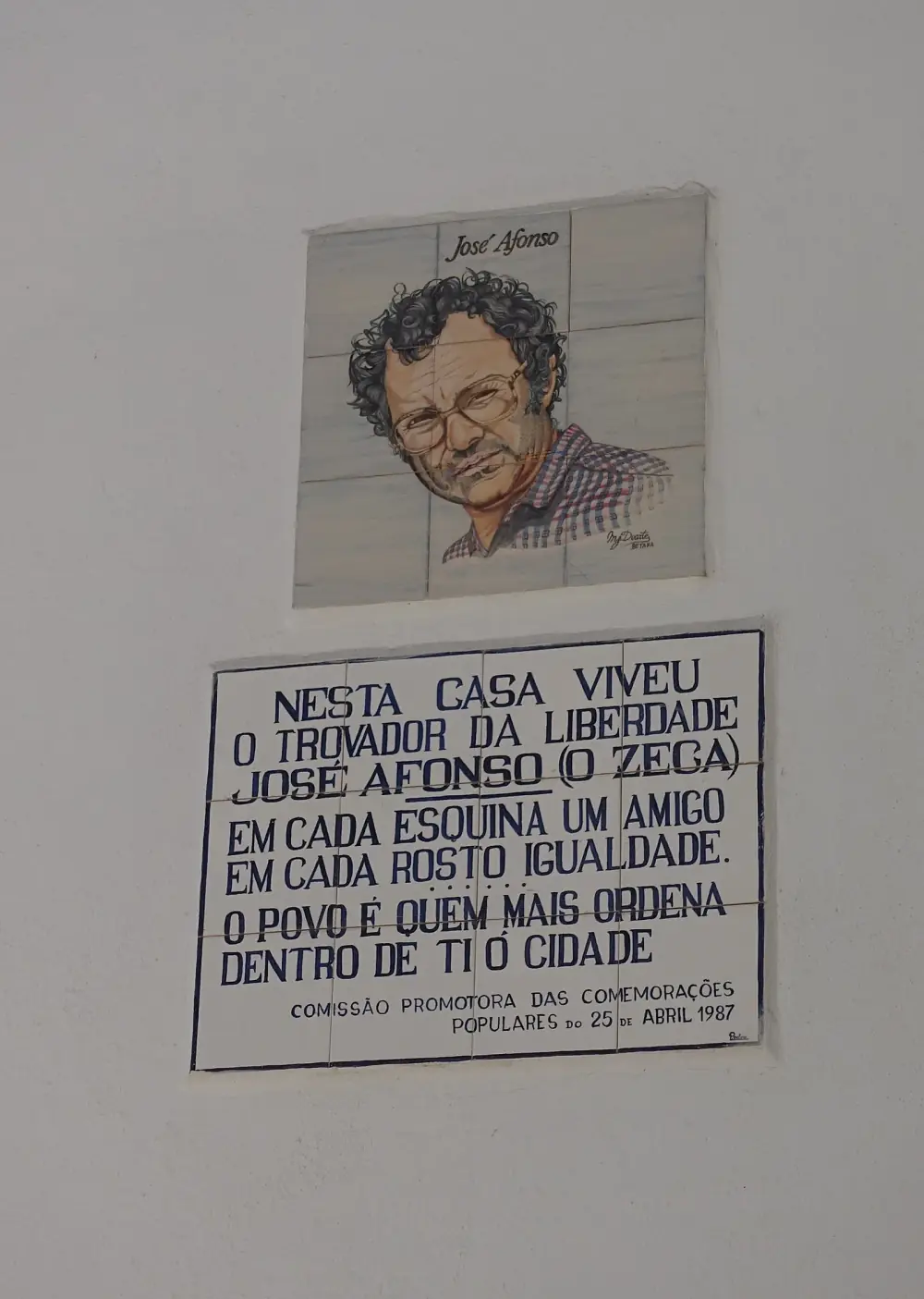 Two tiled panels on a white wall. The upper one is a painted portrait of a man with curly hair, glasses and a checked shirt, titled José Afonso. The lower one, in blue letters, reads that the troubadour of liberty José Afonso, o Zeca, lived in this house, followed by lines from Grândola, Vila Morena.