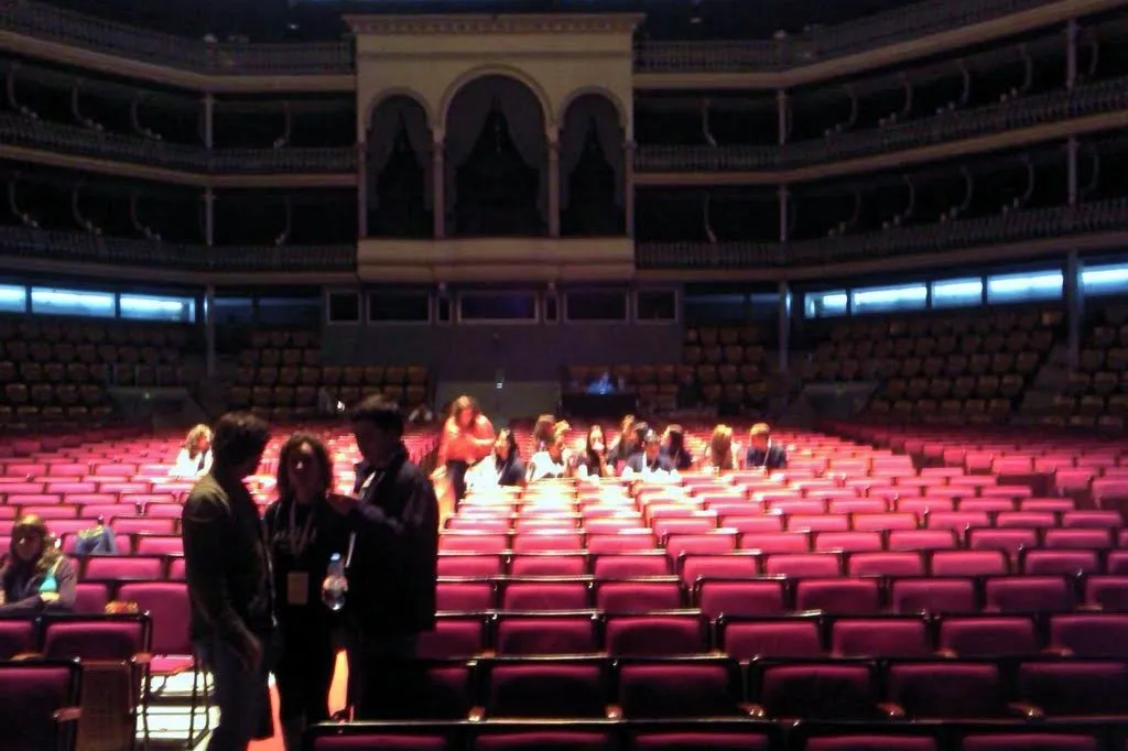 The dim interior of a large round auditorium: rows of red seats in the stalls, a few people standing and sitting among them, and several tiers of balconies curving around the hall above.
