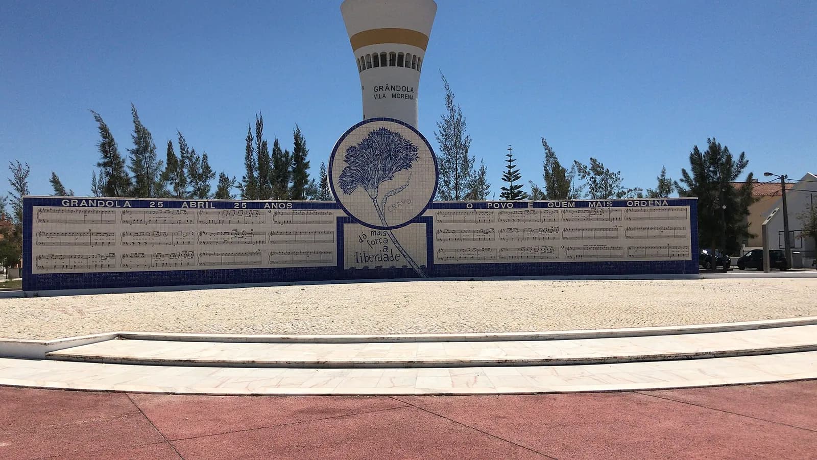 A long wall of blue and white tiles on a paved square, covered with lines of printed music. Lettering along the top reads Grândola 25 Abril 25 anos and O povo é quem mais ordena. In the middle a round panel shows a blue carnation. Behind it rises a white water tower painted with the words Grândola Vila Morena.